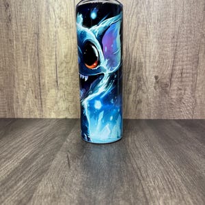 Pu&ograve; includere: Un bicchiere in acciaio inossidabile con una vivace illustrazione di un personaggio alieno blu con grandi occhi e denti affilati. Il design incorpora sfumature di blu, viola e bianco su uno sfondo blu scuro. Il bicchiere ha un coperchio nero e una cannuccia.