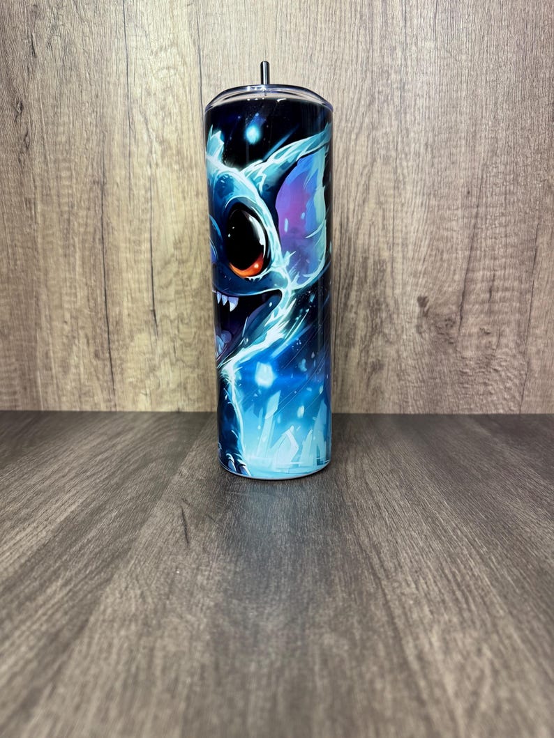 Pu&ograve; includere: Un bicchiere in acciaio inossidabile con una vivace illustrazione di un personaggio alieno blu con grandi occhi e denti affilati. Il design incorpora sfumature di blu, viola e bianco su uno sfondo blu scuro. Il bicchiere ha un coperchio nero e una cannuccia.