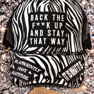 May include: A black and white zebra print trucker hat with the text "BACK THE F**K UP AND STAY THAT WAY." Additional patches read "SO, APPARENTLY I HAVE AN ATTITUDE" and "IT IS WHAT IT IS."
