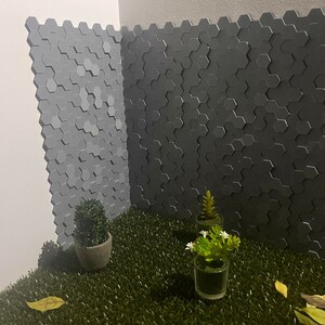 Puede incluir: Primer plano de una pared de azulejos hexagonales grises con tres pequeñas plantas en maceta delante. Las plantas están sobre un lecho de hierba artificial verde.