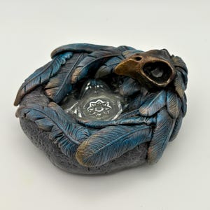 May include: A decorative, round, dark grey trinket dish with blue and bronze-coloured feathers. The dish features a bronze-coloured bird skull and a clear glass insert with a floral design. The feathers are arranged around the dish.