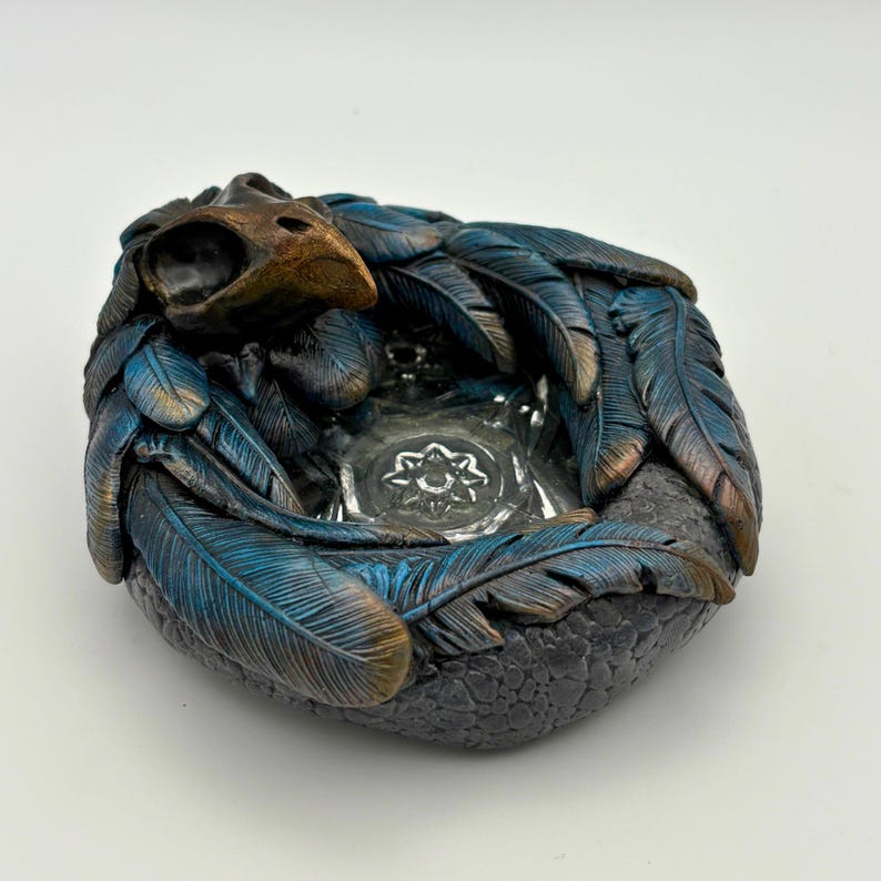 May include: An ornamental ashtray with a bird skull and blue feathers with bronze highlights. The ashtray has a textured, dark grey base and a clear glass centre with a floral design.