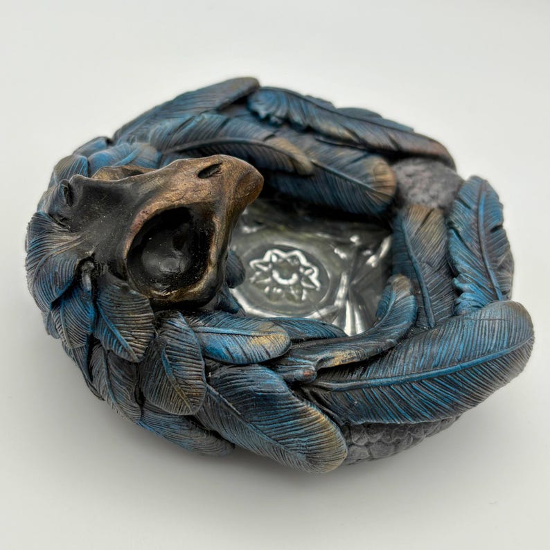 May include: A circular sculpture with a bird-like head and open beak, surrounded by blue and gold-tipped feathers. The sculpture has a dark, weathered appearance and a central, clear glass dish with a floral design.