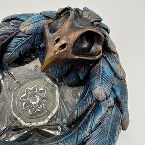 May include: A detailed, close-up of a decorative object featuring a bird skull and feather design. The skull is bronze-coloured, and the feathers are blue and teal. The object has a clear, geometric base with a star-like design.