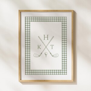 May include: A framed print with a green and white gingham border. The print features a pair of crossed golf clubs with a golf ball in the center. The letters "HKT" are above the clubs.