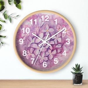 May include: Round wall clock with a light wood frame. The clock face features a watercolor painting of purple and white flowers. White numerals mark the hours, and white hands indicate the time.