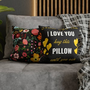 Puede incluir: Dos cojines decorativos sobre un sofá gris. Uno presenta un estampado floral con flores rosas, amarillas y rojas sobre un fondo negro. El otro cojín tiene el texto "I LOVE YOU hug this PILLOW until you can" con corazones amarillos.