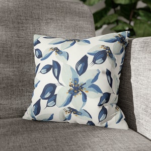 Puede incluir: Cojín cuadrado con fondo blanco y un gran estampado floral en tonos azules y dorados. El cojín está colocado sobre una silla tapizada gris, lo que sugiere un artículo de decoración del hogar.