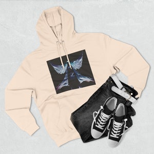 Puede incluir: Sudadera con capucha de color melocotón claro con un gráfico de una figura alada sobre un fondo negro. Se combina con vaqueros oscuros, un cinturón blanco y zapatillas deportivas blancas y negras. Un conjunto casual y elegante.