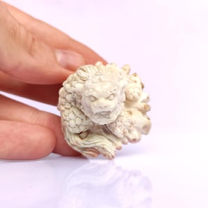 May include: A detailed, off-white sculpture of a mythical creature, possibly a lion or dragon. The carving features intricate details in the fur and facial features, held against a white background.
