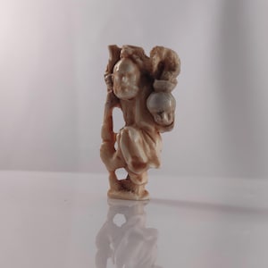 Puede incluir: Una pequeña figurita de marfil tallada de forma intrincada que representa a una persona sosteniendo una calabaza. La figurita está de pie y tiene una expresión facial detallada.