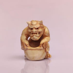Puede incluir: Una figura tallada de una criatura humanoide con cuernos y una expresión feroz, sosteniendo un cuenco. La figura es de color beige claro, con rasgos faciales detallados y una superficie texturizada. El cuenco también es beige y parece estar vacío.