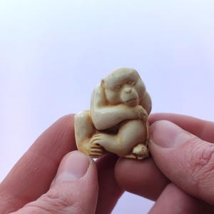 May include: A small, off-white carved figurine of a monkey in a seated position, hugging its knees. The monkey's eyes are closed, suggesting sleep or contemplation. The carving has smooth, rounded features and is held between two hands.