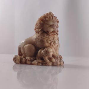May include: A tan-colored lion statue with detailed carving. The lion is seated with a cub at its feet. The statue has a textured surface and is set against a white background. The lion has a detailed mane and facial features.