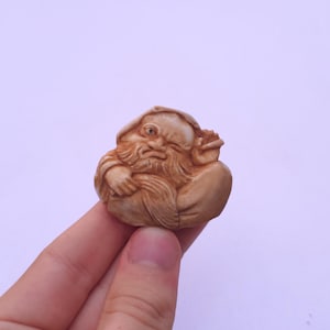 May include: A small, light brown carved figurine depicting a bearded figure with a hood, featuring detailed facial expressions, a prominent beard, and draped clothing. The carving shows intricate textures and lines throughout the surface.