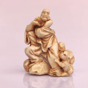 May include: Ivory-colored figurine depicting a seated, bearded figure in flowing robes, possibly a sage or deity. A smaller figure stands nearby, suggesting a narrative or symbolic scene. The sculpture has a detailed, aged appearance.
