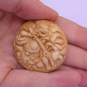 Könnte beinhalten: Ein kreisförmiger, hellbrauner, geschnitzter Anhänger mit einem Reliefdesign einer lächelnden Figur in fließenden Gewändern, die ein Schwert hält. Die Schnitzerei zeigt detaillierte Gesichtszüge, die geschwungene Klinge und wirbelnde Stoffmuster.