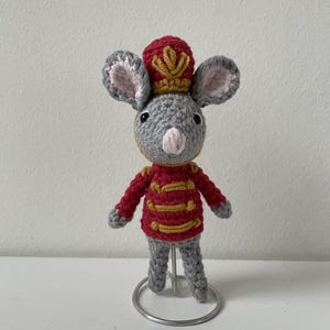 May include: A handmade crochet mouse figurine wearing a red and gold military-style jacket and hat. The gray mouse has large ears with pink inner ears and a pink nose. The figurine is standing on a silver metal stand.