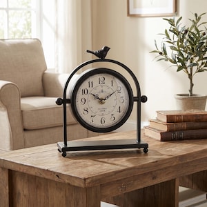 May include: A black metal table clock featuring a circular face with an off-white background, black Arabic numerals, and the text Old Town Clock. The clock is mounted on a stand with a small bird figurine on top and rests on a rectangular base.
