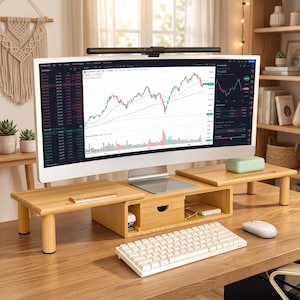 May include: A light-colored bamboo monitor stand with a central drawer and two side platforms, supporting a curved computer monitor displaying financial charts. A white wireless keyboard and mouse sit on the desk surface in front of the stand.