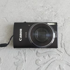 Puede incluir: Una cámara digital Canon IXUS negra que presenta un prominente barril de lente circular con el texto 10x optical zoom, una unidad de flash integrada y una pequeña correa de muñeca negra unida al lado del cuerpo rectangular.