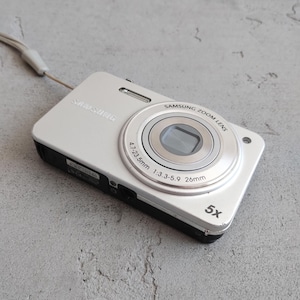 May include: A silver Samsung digital camera with a black base and a wrist strap. The lens barrel is labeled Samsung Zoom Lens 4.7-23.5mm 1:3.3-5.9 26mm and features a 5x optical zoom indicator on the front face of the camera body.