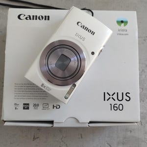 May include: A white Canon IXUS 160 digital camera with an 8x optical zoom lens, resting on its original white product box. The box features the Canon logo, IXUS 160 branding, and technical specifications including 20.0 megapixels and 6.8 cm LCD screen.