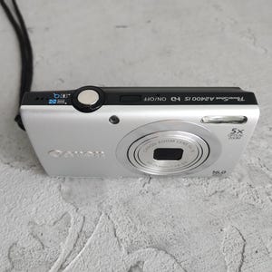 Puede incluir: Una cámara digital Canon PowerShot A2400 IS de color plateado con un panel superior negro. La cámara cuenta con un objetivo de zoom óptico de 5x, un sensor de 16,0 megapíxeles, un flash y una correa de muñeca negra unida al lateral.