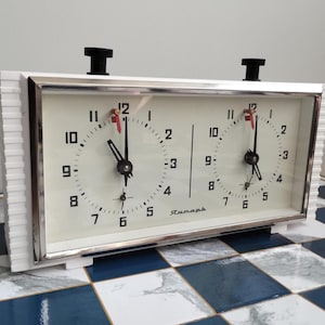 May include: A vintage white chess clock with two clock faces, black hands, and a red second hand. The clock has a silver frame and black knobs on top. The clock is set on a blue and white checkered surface.