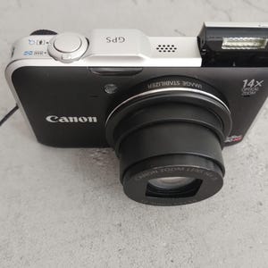 Puede incluir: Una cámara digital Canon PowerShot negra y plateada con un objetivo de zoom óptico de 14x. El panel superior tiene un acabado plateado con un botón disparador y la etiqueta GPS. La cámara incluye un flash emergente y una correa de muñeca negra.