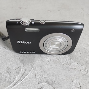 May include: A black Nikon Coolpix digital camera featuring a silver lens barrel with text reading NIKKOR 5X WIDE OPTICAL ZOOM 4.6-23.0mm 1:3.2-6.5. The camera includes a top-mounted silver shutter button, zoom toggle, and a black wrist strap attached to the side.