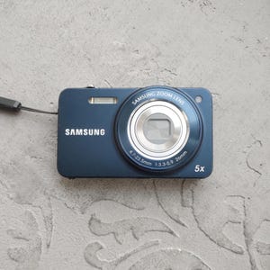 May include: A dark blue Samsung digital camera featuring a Samsung Zoom Lens with 4.7-23.5mm 1:3.3-5.9 26mm specifications and 5x zoom capability. The front of the device displays the Samsung brand name and includes a built-in flash unit.