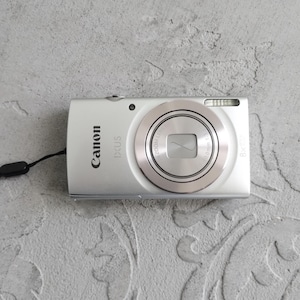 May include: A silver Canon IXUS digital camera featuring an 8x optical zoom lens, a built-in flash, and a black wrist strap. The front of the camera displays the Canon and IXUS brand names, with lens specifications printed around the circular lens housing.