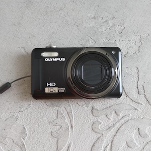 Op de afbeelding: Een zwarte Olympus digitale camera met een 10x groothoek optische zoomlens, een HD-label en een klein polsbandje. De voorzijde van het apparaat toont de merknaam Olympus, de tekst HD en het label 10x Super Wide in het wit.