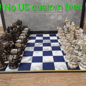 Puede incluir: Un juego de ajedrez con piezas detalladas de estilo antiguo en tonos dorados y bronce. El tablero presenta un patrón de cuadros azules y blancos. Las piezas están dispuestas en el tablero, listas para jugar. El conjunto se muestra sobre una superficie de madera.