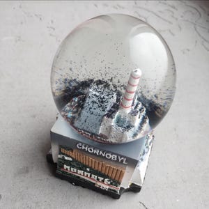 May include: A snow globe depicting the Chernobyl nuclear power plant. The clear glass sphere contains water and black glitter. The base is rectangular and features the word "CHORNOBYL". The miniature plant model includes a red and white striped chimney.