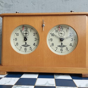 May include: A wooden analog chess clock featuring two circular clock faces with black numerals, black hands, and a small black knight icon on a checkered base. A silver metal lever is positioned at the top center between the two clock faces.