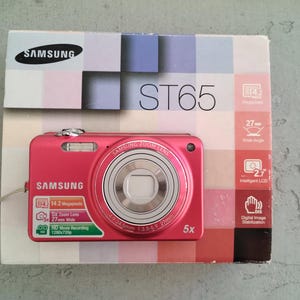 Puede incluir: Una cámara digital Samsung ST65 de color rosa brillante con lente de zoom 5x, colocada sobre su caja original. La caja presenta una cuadrícula de cuadrados pastel, el logotipo de Samsung y especificaciones técnicas como 14,2 megapíxeles y 27 mm.