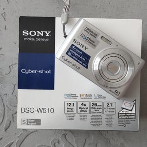 Puede incluir: Una cámara digital Sony Cyber-shot DSC-W510 plateada con correa de muñeca, sobre su caja de producto blanca. La cámara cuenta con zoom óptico de 4x, 12,1 megapíxeles y un objetivo gran angular de 26 mm. La caja muestra las especificaciones técnicas.