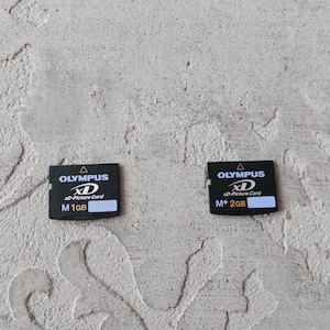 Op de afbeelding: Twee zwarte Olympus xD-Picture Cards. De linkerkaart is gelabeld met M 1GB en de rechterkaart met M+ 2GB. Beide kaarten bevatten witte tekst, het Olympus-logo en een klein wit rechthoekig vlak voor tekst in de rechterbenedenhoek.