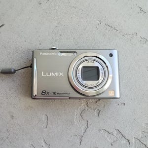 Puede incluir: Una cámara digital Panasonic Lumix de color plateado con zoom óptico de 8x, resolución de 16 megapíxeles y lente Leica DC Vario-Elmar. La parte frontal muestra el nombre de la marca, el modelo y una pequeña correa de muñeca gris.