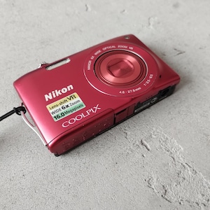 Może przedstawiać: Czerwony aparat cyfrowy Nikon Coolpix z 6-krotnym zoomem optycznym, 16,0 megapikselami i technologią lens-shift VR. Korpus aparatu posiada czarny pasek na nadgarstek przymocowany z boku oraz wyraźnie widoczną nazwę marki i modelu urządzenia.