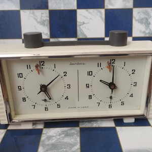 May include: A vintage Jantar chess clock with a cream case and two clock faces. It features a black handle and a silver frame. The clock faces show black hands and Arabic numerals. The text "MADE IN USSR" is visible.