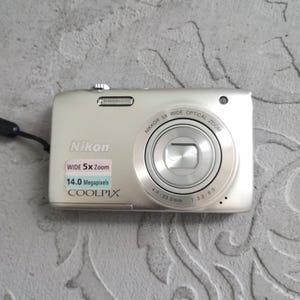 Puede incluir: Una cámara digital Nikon Coolpix plateada con un objetivo zoom óptico gran angular Nikkor 5x, un sensor de 14,0 megapíxeles y un cuerpo rectangular con flash integrado. La parte frontal muestra el texto WIDE 5x Zoom y 14.0 Megapixels.