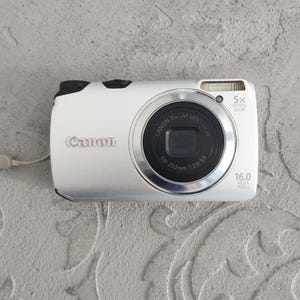 Puede incluir: Una cámara digital Canon plateada con zoom óptico de 5x, 16,0 megapíxeles y un objetivo de 5,0-25,0 mm 1:2,8-5,9. El dispositivo compacto incluye un flash integrado, un botón disparador y un interruptor de encendido en la parte superior.