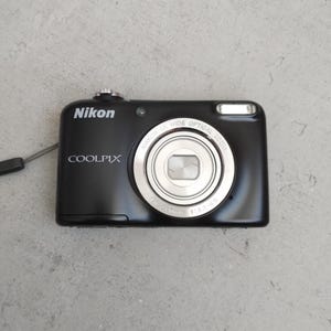 Pode incluir: Uma câmara digital Nikon Coolpix preta com uma objetiva prateada que apresenta o texto NIKKOR 5X WIDE OPTICAL ZOOM 4.6-23.0mm 1:3.2-6.5 impresso à volta da lente. O corpo da câmara inclui um botão de disparo prateado e um flash.