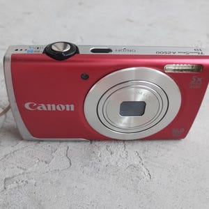 May include: A red Canon PowerShot A2500 digital camera with a silver lens housing, a black shutter button, and an on/off switch. The front features the Canon logo, a flash, and text indicating 5x optical zoom and 16.0 megapixels.