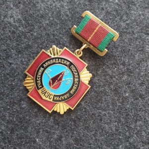 Puede incluir: Una medalla de metal dorado con forma de cruz de esmalte rojo y un círculo central azul claro. El centro muestra un símbolo de gota roja y texto en cirílico. Está sujeta a una barra de cinta rectangular con rayas verdes y rojas.