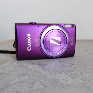 May include: A purple Canon IXUS digital camera featuring a 12x optical zoom lens, a built-in flash, and a black wrist strap. The front of the camera displays the Canon logo, the IXUS model name, and technical specifications printed on the lens ring.