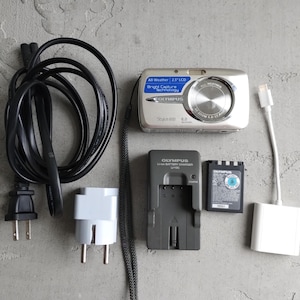 Peut inclure: Un appareil photo numérique argenté Olympus Stylus 600 avec dragonne, un cordon d'alimentation noir, un adaptateur mural blanc, un chargeur de batterie gris Olympus Li-ion, une batterie noire et un câble adaptateur Lightning vers USB.